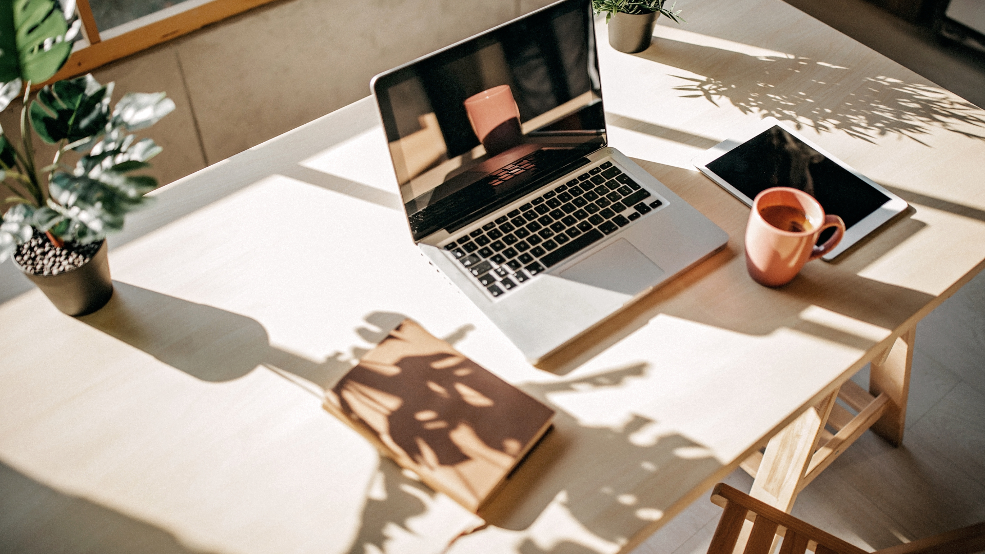 A sunlit desk with a laptop, tablet, notebook, and a cup of tea